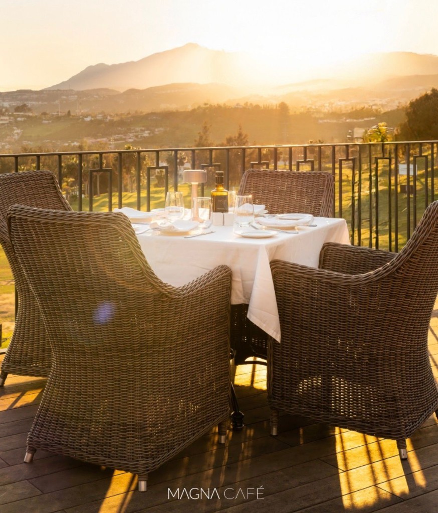 Magna Café terrace at sunset with a set table for four, wicker armchairs and panoramic view of Marbella mountains